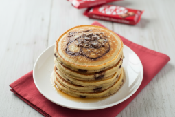 Un généreux stack de pancakes dorés est servi sur une assiette, avec des éclats de chocolat fondants sur le dessus. En arrière‑plan, on aperçoit des barres de chocolat emballées ajoutant une touche gourmande à la scène.